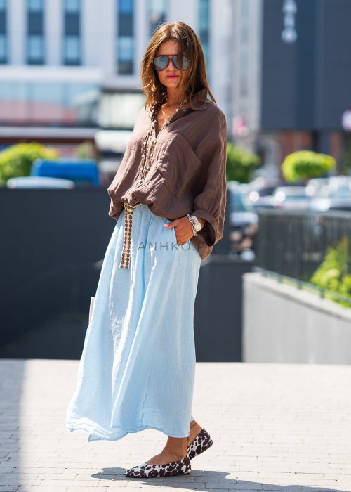 Brązowa koszula lniana damska o luźnym kroju z kołnierzykiem i kieszonką na piersi, zestawiona z jasną, lnianą spódnicą i dodatkami w naturalnym stylu.