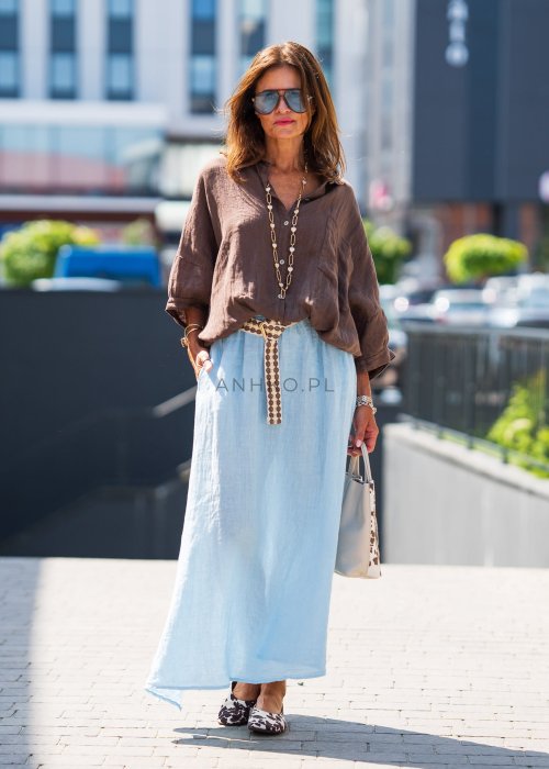 Brązowa koszula lniana damska o luźnym kroju z kołnierzykiem i kieszonką na piersi, zestawiona z jasną, lnianą spódnicą i dodatkami w naturalnym stylu.