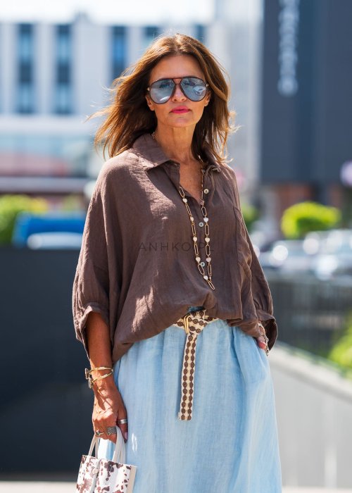 Brązowa koszula lniana damska o luźnym kroju z kołnierzykiem i kieszonką na piersi, zestawiona z jasną, lnianą spódnicą i dodatkami w naturalnym stylu.