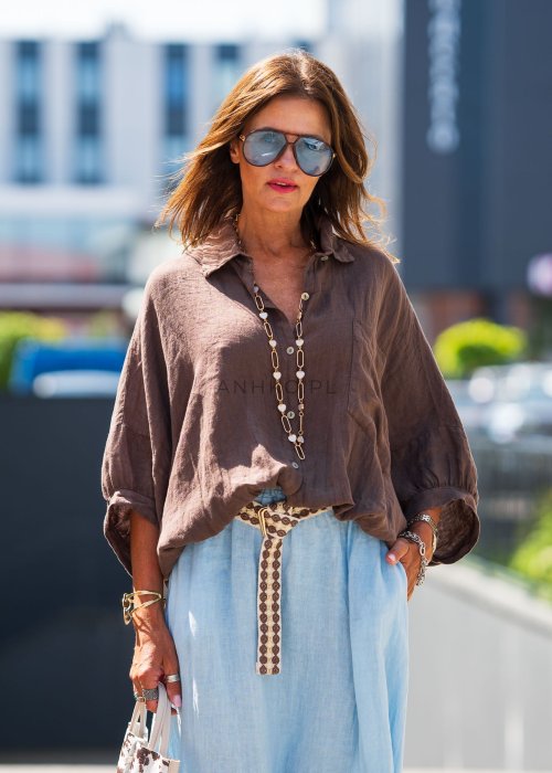 Brązowa koszula lniana damska o luźnym kroju z kołnierzykiem i kieszonką na piersi, zestawiona z jasną, lnianą spódnicą i dodatkami w naturalnym stylu.