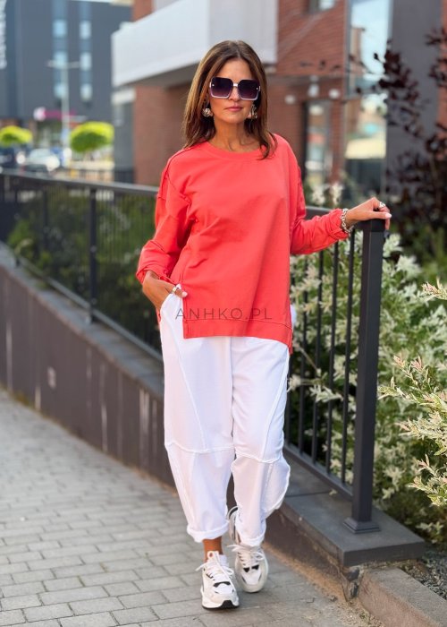Damska bluza oversize w intensywnym koralowym kolorze z długim rękawem i okrągłym dekoltem. Stylizacja miejska zestawiona z jasnymi spodniami i dużymi okularami przeciwsłonecznymi.