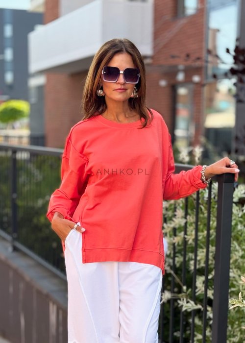 Damska bluza oversize w intensywnym koralowym kolorze z długim rękawem i okrągłym dekoltem. Stylizacja miejska zestawiona z jasnymi spodniami i dużymi okularami przeciwsłonecznymi.