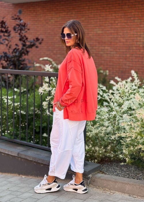 Damska bluza oversize w intensywnym koralowym kolorze z długim rękawem i okrągłym dekoltem. Stylizacja miejska zestawiona z jasnymi spodniami i dużymi okularami przeciwsłonecznymi.