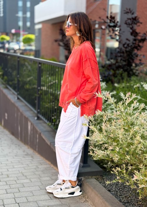 Damska bluza oversize w intensywnym koralowym kolorze z długim rękawem i okrągłym dekoltem. Stylizacja miejska zestawiona z jasnymi spodniami i dużymi okularami przeciwsłonecznymi.