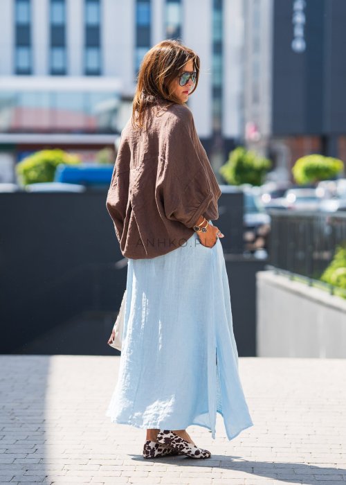 Brązowa koszula lniana damska o luźnym kroju z kołnierzykiem i kieszonką na piersi, zestawiona z jasną, lnianą spódnicą i dodatkami w naturalnym stylu.