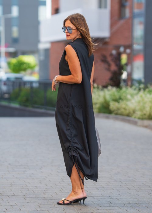 Czarna sukienka maxi bez rękawów ze stójką i regulowanym dołem na troczki. Modelka prezentuje minimalistyczną miejską stylizację z sandałami i torebką.