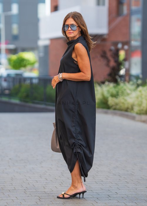 Czarna sukienka maxi bez rękawów ze stójką i regulowanym dołem na troczki. Modelka prezentuje minimalistyczną miejską stylizację z sandałami i torebką.