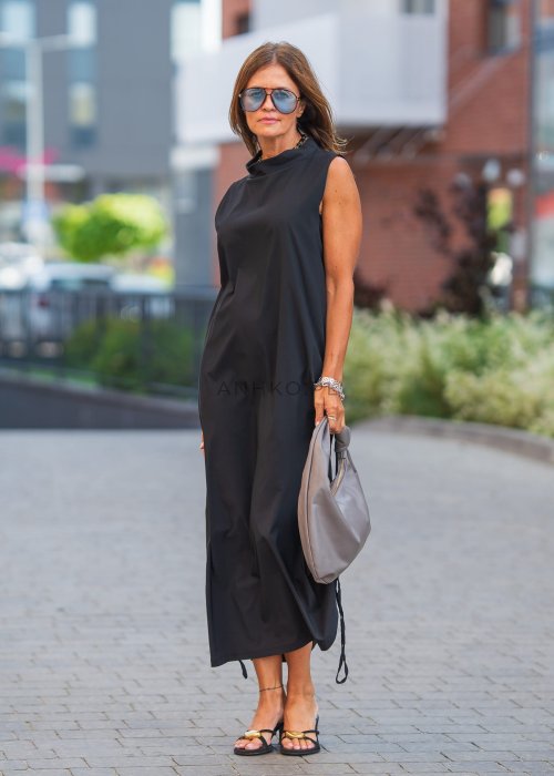 Czarna sukienka maxi bez rękawów ze stójką i regulowanym dołem na troczki. Modelka prezentuje minimalistyczną miejską stylizację z sandałami i torebką.