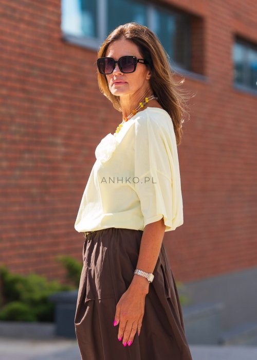 Żółta bluzka z krótkim rękawem o luźnym kroju, ozdobiona materiałowym kwiatem, zestawiona z brązowymi spodniami z wysokim stanem