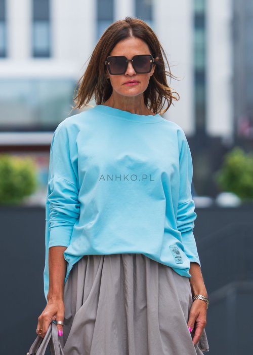 Bluza damska oversize w kolorze baby blue z długim rękawem i naszywką na froncie. Luźny fason w stylizacji z szeroką spódnicą i dodatkami w miejskim stylu.