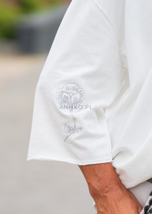 Biała bluzka oversize damska z szerokim dekoltem i haftowanym detalem na rękawie, zestawiona ze spodniami moro w casualowej stylizacji miejskiej.