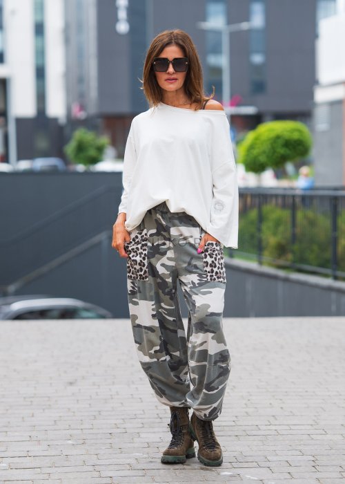 Biała bluzka oversize damska z szerokim dekoltem i haftowanym detalem na rękawie, zestawiona ze spodniami moro w casualowej stylizacji miejskiej.