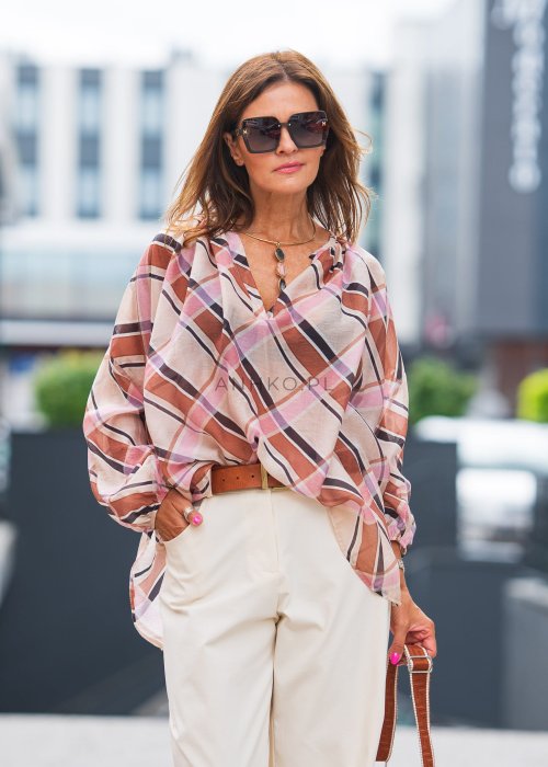 Wzorzysta bluzka w różowo-brązową kratę, kremowe spodnie i brązowy pasek, do tego duże okulary i delikatny naszyjnik.