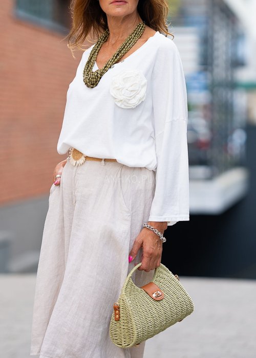 Biała bluzka z długim rękawem, o luźnym kroju, z dekoltem w serek ozdobionym dużym kwiatem z tkaniny po lewej stronie.