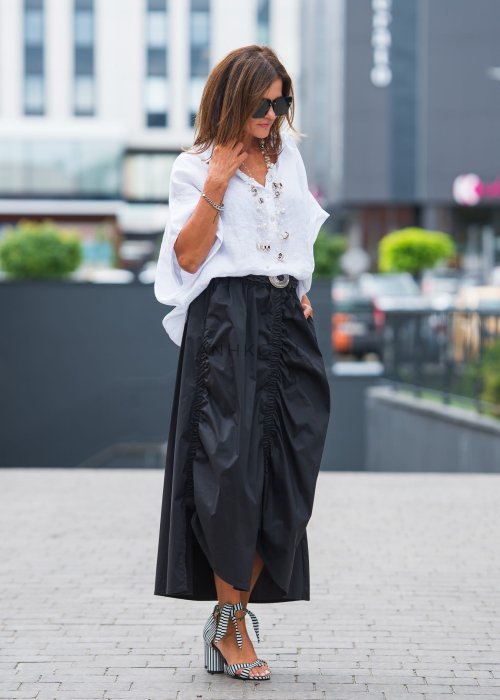 Biała, luźna koszula z kołnierzykiem, czarne spodnie z ozdobnym paskiem i długi naszyjnik z perełkami.