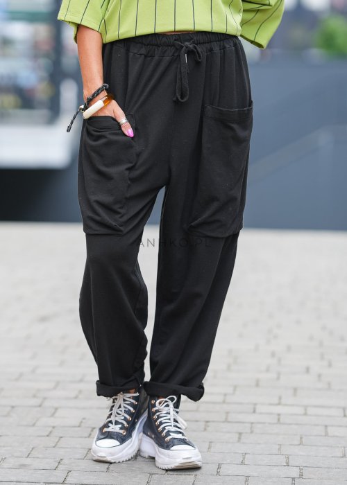 Czarne spodnie damskie o luźnym kroju z elastycznym pasem i dużymi naszywanymi kieszeniami z przodu. Model zestawiony ze sneakersami i zieloną bluzą w paski w miejskiej stylizacji