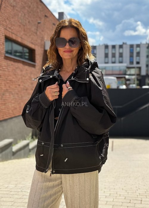 Czarna kurtka damska oversize z kapturem i kontrastowymi przeszyciami, pokazana w miejskiej stylizacji. Model zapinany na suwak, o luźnym kroju, zestawiony z jasnymi spodniami.