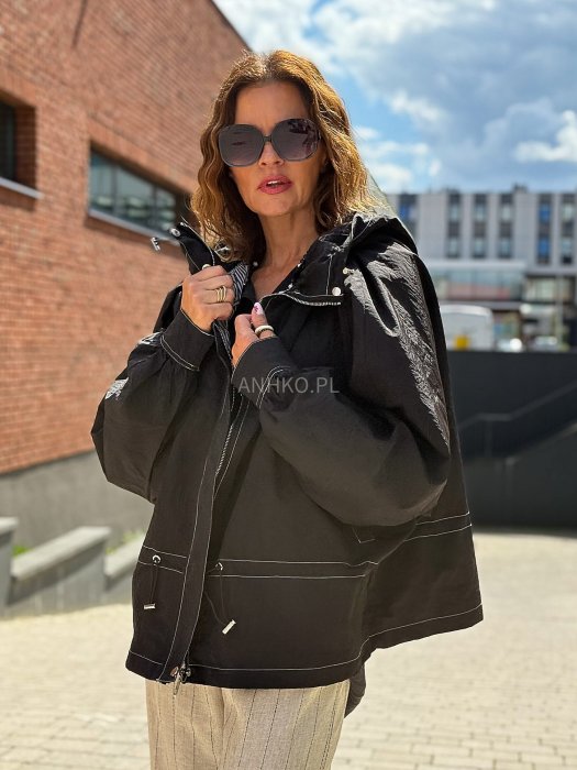Czarna kurtka damska oversize z kapturem i kontrastowymi przeszyciami, pokazana w miejskiej stylizacji. Model zapinany na suwak, o luźnym kroju, zestawiony z jasnymi spodniami.