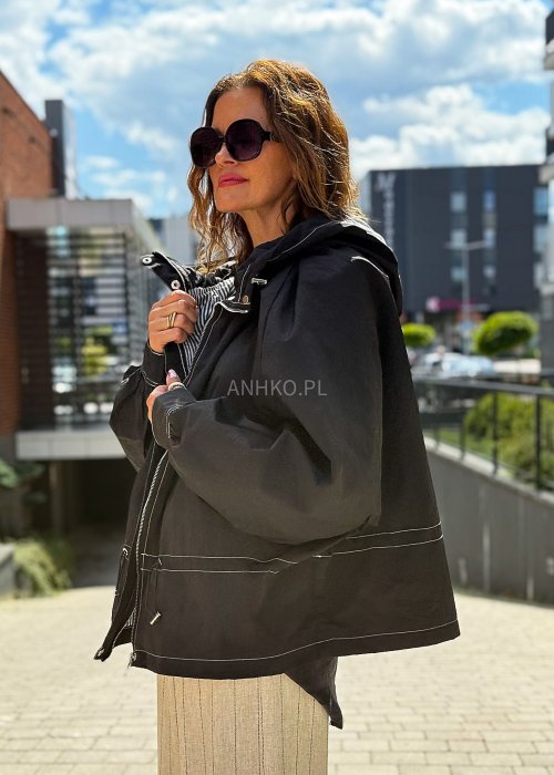 Czarna kurtka damska oversize z kapturem i kontrastowymi przeszyciami, pokazana w miejskiej stylizacji. Model zapinany na suwak, o luźnym kroju, zestawiony z jasnymi spodniami.