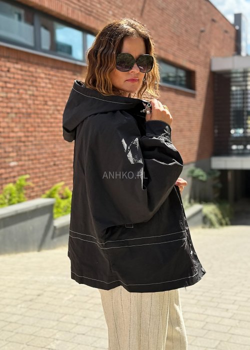 Czarna kurtka damska oversize z kapturem i kontrastowymi przeszyciami, pokazana w miejskiej stylizacji. Model zapinany na suwak, o luźnym kroju, zestawiony z jasnymi spodniami.