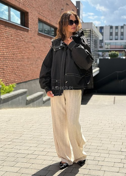 Czarna kurtka damska oversize z kapturem i kontrastowymi przeszyciami, pokazana w miejskiej stylizacji. Model zapinany na suwak, o luźnym kroju, zestawiony z jasnymi spodniami.