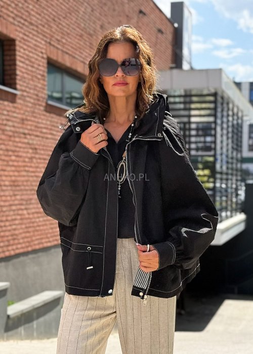Czarna kurtka damska oversize z kapturem i kontrastowymi przeszyciami, pokazana w miejskiej stylizacji. Model zapinany na suwak, o luźnym kroju, zestawiony z jasnymi spodniami.