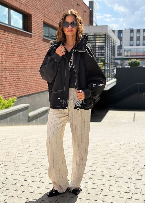 Czarna kurtka damska oversize z kapturem i kontrastowymi przeszyciami, pokazana w miejskiej stylizacji. Model zapinany na suwak, o luźnym kroju, zestawiony z jasnymi spodniami.