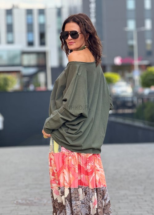 Khaki bluzka damska oversize z długim rękawem i dekoltem V, stylizowana lekko opuszczona na ramię. Model prezentowany ze wzorzystą spódnicą midi i naszyjnikiem w zielonym kolorze.