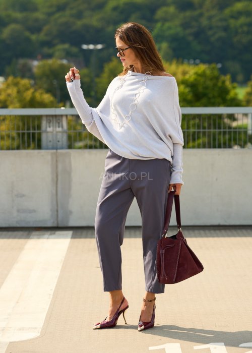 Jasnoszary sweter oversize o nietoperzowym kroju z dekoltem łódka i asymetrycznym dołem zestawiony z grafitowymi eleganckimi spodniami oraz dużymi okularami przeciwsłonecznymi.
