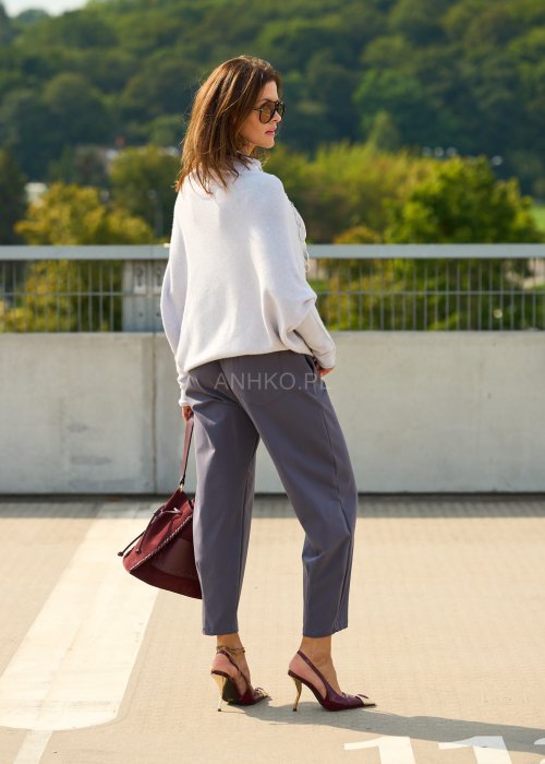 Jasnoszary sweter oversize o nietoperzowym kroju z dekoltem łódka i asymetrycznym dołem zestawiony z grafitowymi eleganckimi spodniami oraz dużymi okularami przeciwsłonecznymi.