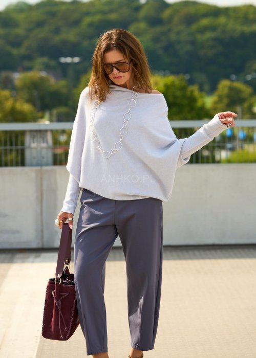 Jasnoszary sweter oversize o nietoperzowym kroju z dekoltem łódka i asymetrycznym dołem zestawiony z grafitowymi eleganckimi spodniami oraz dużymi okularami przeciwsłonecznymi.