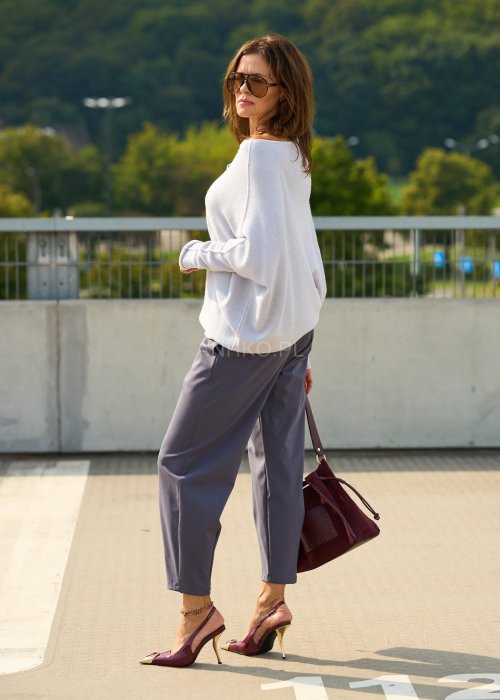 Jasnoszary sweter oversize o nietoperzowym kroju z dekoltem łódka i asymetrycznym dołem zestawiony z grafitowymi eleganckimi spodniami oraz dużymi okularami przeciwsłonecznymi.