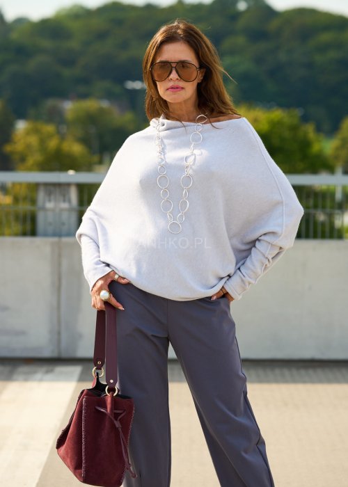 Jasnoszary sweter oversize o nietoperzowym kroju z dekoltem łódka i asymetrycznym dołem zestawiony z grafitowymi eleganckimi spodniami oraz dużymi okularami przeciwsłonecznymi.