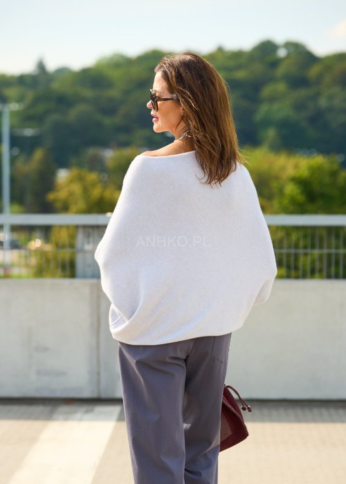 Jasnoszary sweter oversize o nietoperzowym kroju z dekoltem łódka i asymetrycznym dołem zestawiony z grafitowymi eleganckimi spodniami oraz dużymi okularami przeciwsłonecznymi.