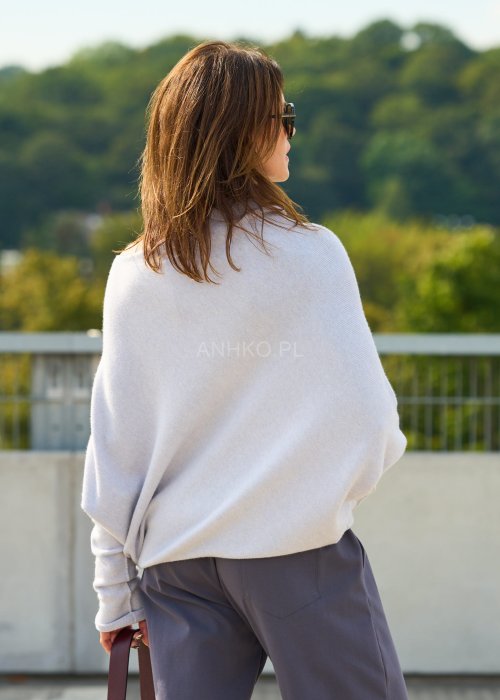 Jasnoszary sweter oversize o nietoperzowym kroju z dekoltem łódka i asymetrycznym dołem zestawiony z grafitowymi eleganckimi spodniami oraz dużymi okularami przeciwsłonecznymi.