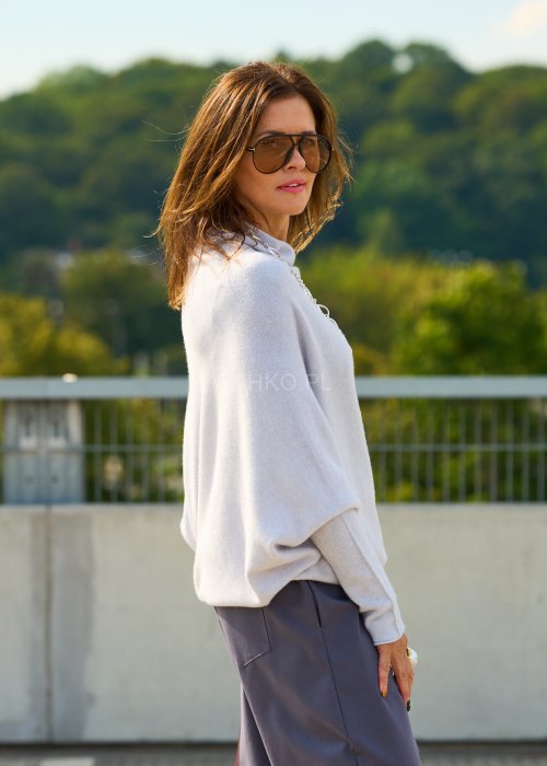 Jasnoszary sweter oversize o nietoperzowym kroju z dekoltem łódka i asymetrycznym dołem zestawiony z grafitowymi eleganckimi spodniami oraz dużymi okularami przeciwsłonecznymi.