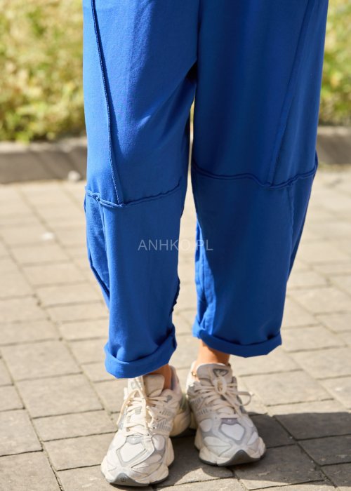 Niebieskie damskie spodnie dresowe o luźnym kroju z ozdobnymi przeszyciami i wiązaniem w pasie. Model z kieszeniami, pokazany w codziennej stylizacji ze sneakersami.