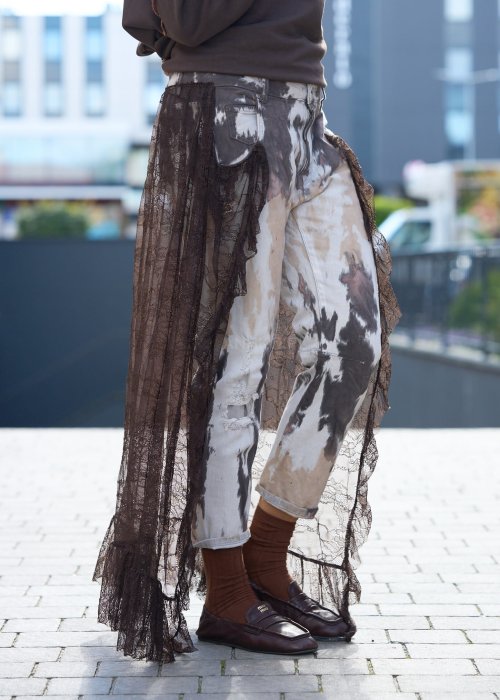 Damskie spodnie w odcieniach brązu i beżu, z nałożoną koronkową warstwą z ciemnobrązowej siateczki, stylizowane z brązowymi mokasynami i skarpetkami.