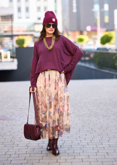 Śliwkowa bluza damska oversize z długim rękawem i naszywką, zestawiona ze wzorzystą spódnicą w stylizacji miejskiej.