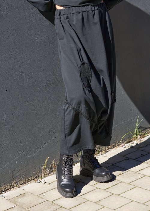 Czarne spodnie damskie oversize z obniżonym krokiem i ozdobnymi aplikacjami, zestawione z botkami i luźną bluzą w nowoczesnej stylizacji streetwear.