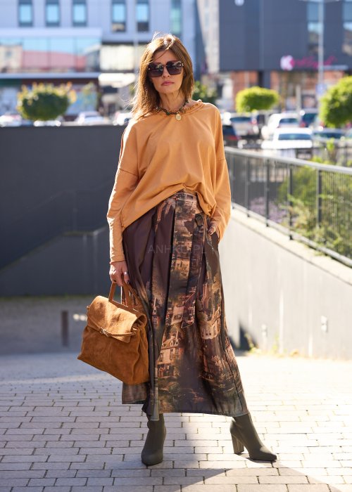 Karmelowa torebka z zamszu o miękkiej strukturze, z klapą i metalowym zapięciem, trzymana w dłoni kobiety w jesiennej stylizacji z długą, wzorzystą spódnicą.