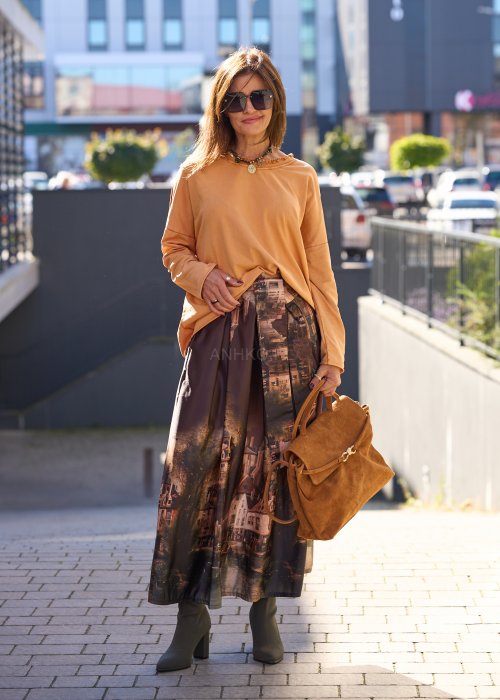 Karmelowa torebka z zamszu o miękkiej strukturze, z klapą i metalowym zapięciem, trzymana w dłoni kobiety w jesiennej stylizacji z długą, wzorzystą spódnicą.