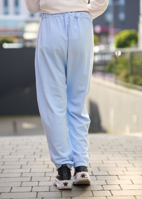 Jasnoniebieskie spodnie harem damskie z dużymi kieszeniami ozdobionymi perłami i elastyczną talią, stylizowane w miejskim looku ze sneakersami.