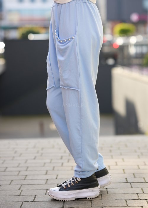 Jasnoniebieskie spodnie harem damskie z dużymi kieszeniami ozdobionymi perłami i elastyczną talią, stylizowane w miejskim looku ze sneakersami.