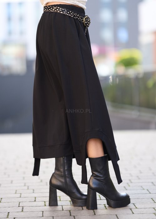 Czarna spódnica dzianinowa o asymetrycznym dole i elastycznym pasie z ozdobnym paskiem. Model pokazany w stylizacji z jasnym swetrem i czarnymi botkami na obcasie.