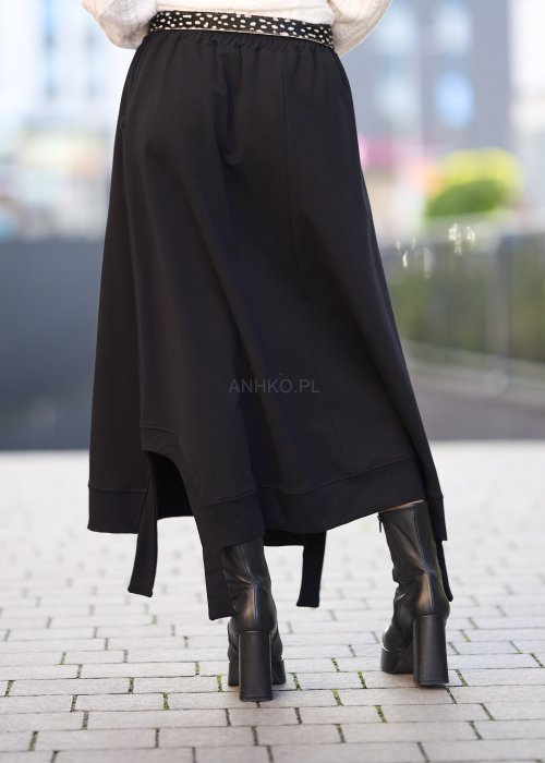 Czarna spódnica dzianinowa o asymetrycznym dole i elastycznym pasie z ozdobnym paskiem. Model pokazany w stylizacji z jasnym swetrem i czarnymi botkami na obcasie.