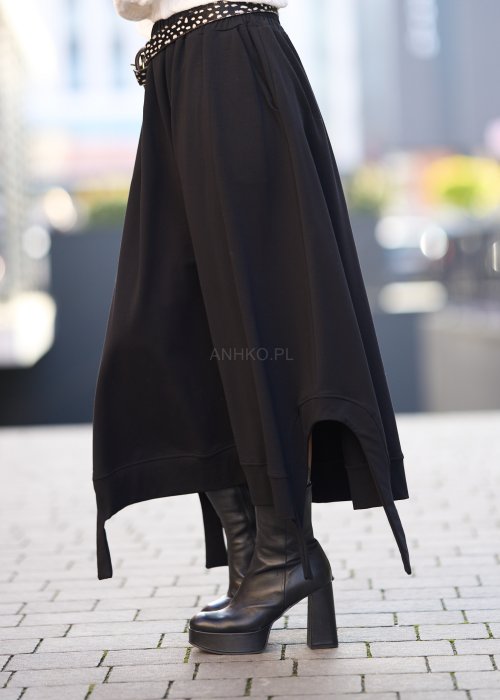 Czarna spódnica dzianinowa o asymetrycznym dole i elastycznym pasie z ozdobnym paskiem. Model pokazany w stylizacji z jasnym swetrem i czarnymi botkami na obcasie.