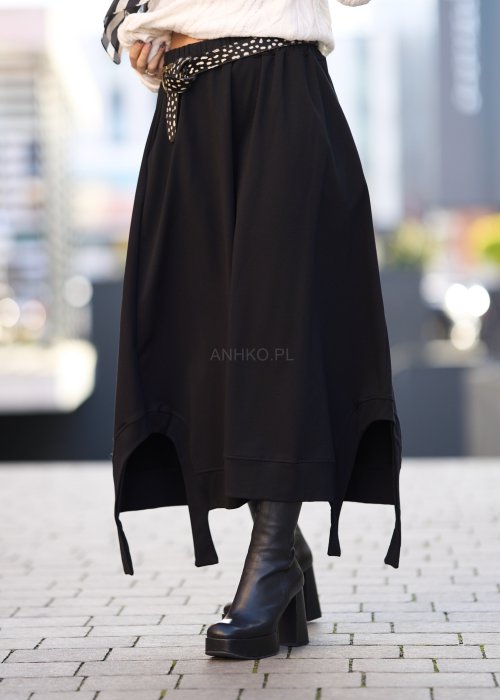 Czarna spódnica dzianinowa o asymetrycznym dole i elastycznym pasie z ozdobnym paskiem. Model pokazany w stylizacji z jasnym swetrem i czarnymi botkami na obcasie.