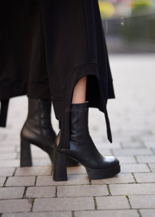 Czarna spódnica dzianinowa o asymetrycznym dole i elastycznym pasie z ozdobnym paskiem. Model pokazany w stylizacji z jasnym swetrem i czarnymi botkami na obcasie.