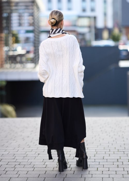 Czarna spódnica dzianinowa o asymetrycznym dole i elastycznym pasie z ozdobnym paskiem. Model pokazany w stylizacji z jasnym swetrem i czarnymi botkami na obcasie.